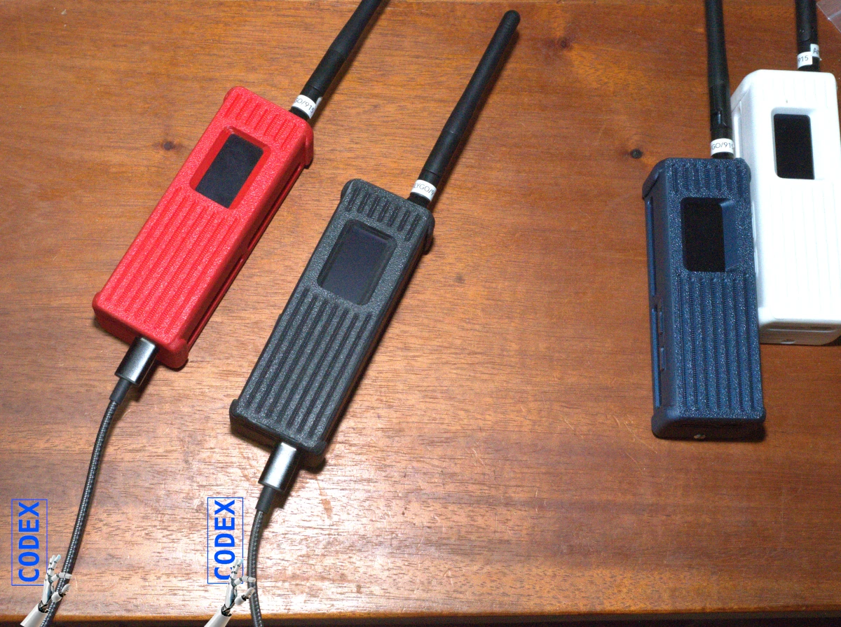 four portable radio units on a desktop, two are connected with USB cable and there are robotic hands with labels of CODEX reaching upo from the edge of the photo towards the units connected by USB